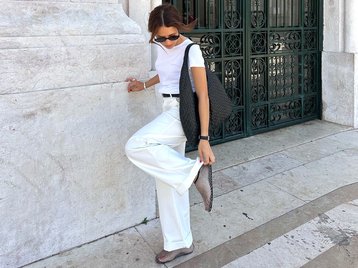 fashion influencer Debora Rosa poses outside a marble wall in Portugal wearing retro black oval sunglasses, white T-shirt, black woven hobo tote bag, black belt, white relaxed pants, and crystal embellished black mesh flats