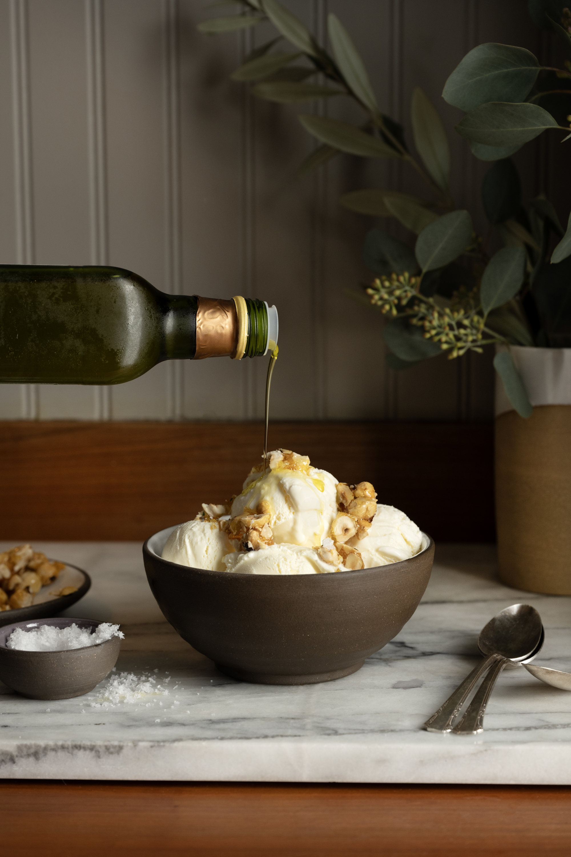 Image of a dark brown, ceramic bowl on a white marble countertop. The bowl is filled with vanilla ice cream covered in hazelnuts, with a bottle of olive oil being poured over it.