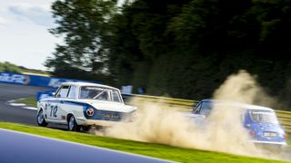 A white and blue classic touring car drifts on a racetrack, kicking up dust as it competes with a blue car in the background