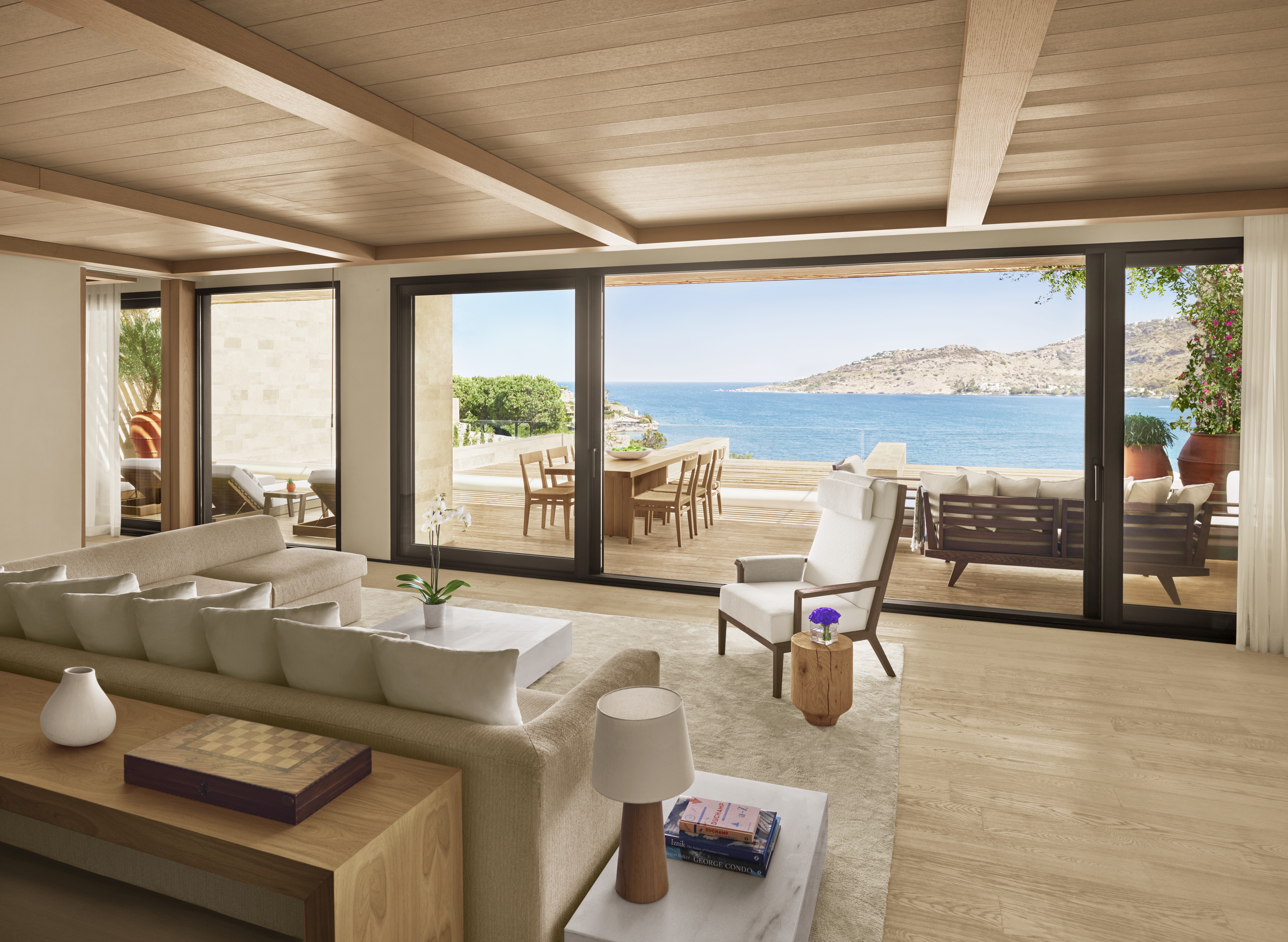 A creme-tinted resort room features wooden furnishings mixed with beige and white fabrics and a sprawling view over the Agean sea.