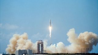 a white and black rocket lifts off into a blue sky leaving behind a plume spread across its launch site