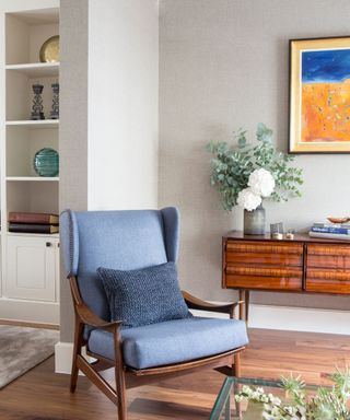 A blue and wooden armchair in a tidy living room with a wooden sideboard, vase of flowers, stack of coffee table books and framed bright artwork with open shelves to the left seen in the next room