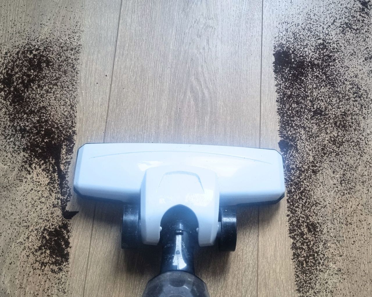 A wooden floor with coffee grounds being suctioned up by a white vacuum