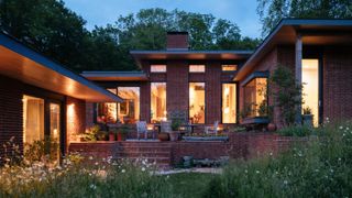 evening shot of low level brick house exterior taken from rear of house showing windows lit up and a patio area