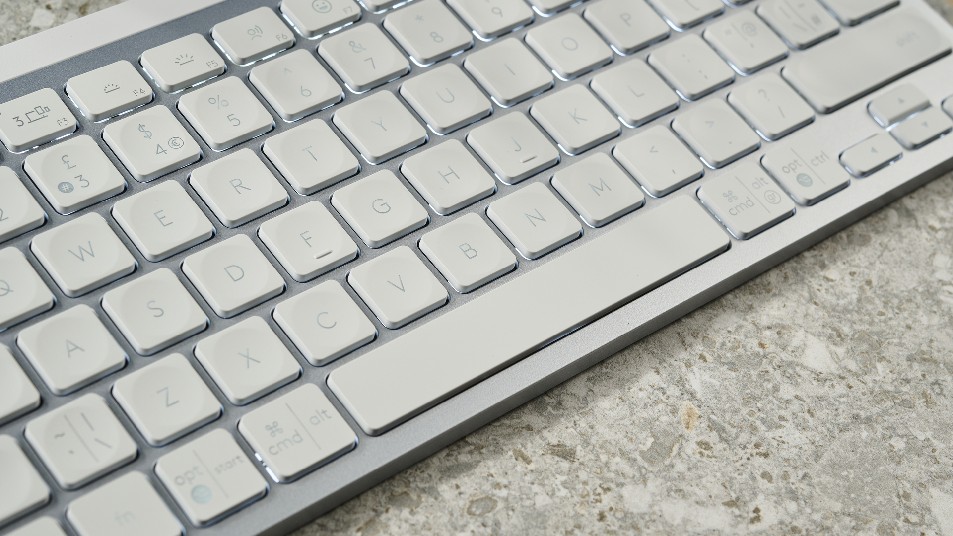 A photograph of the Logitech MX Keys Mini in light gray, positioned on a stone slab with a blue wall in the background.