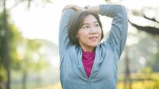 woman wearing a light blue zipper exercise top stretching her upper arm behind her facing the camera but looking away. she's in a park setting with a background of trees blurred behind her.