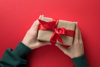 A person's hands holding a brown paper wrapped gift with red ribbon on, in front of a red background.