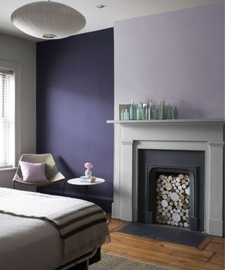 A dark purple and lilac bedroom with a black fireplace, a beige bed, and a table and chair set with blush pink accessories – by Benjamin Moore using Benjamin Moore Aura Matte Dark Lilac 2070- 30, Aura Matte Lavender Mist 2070-60