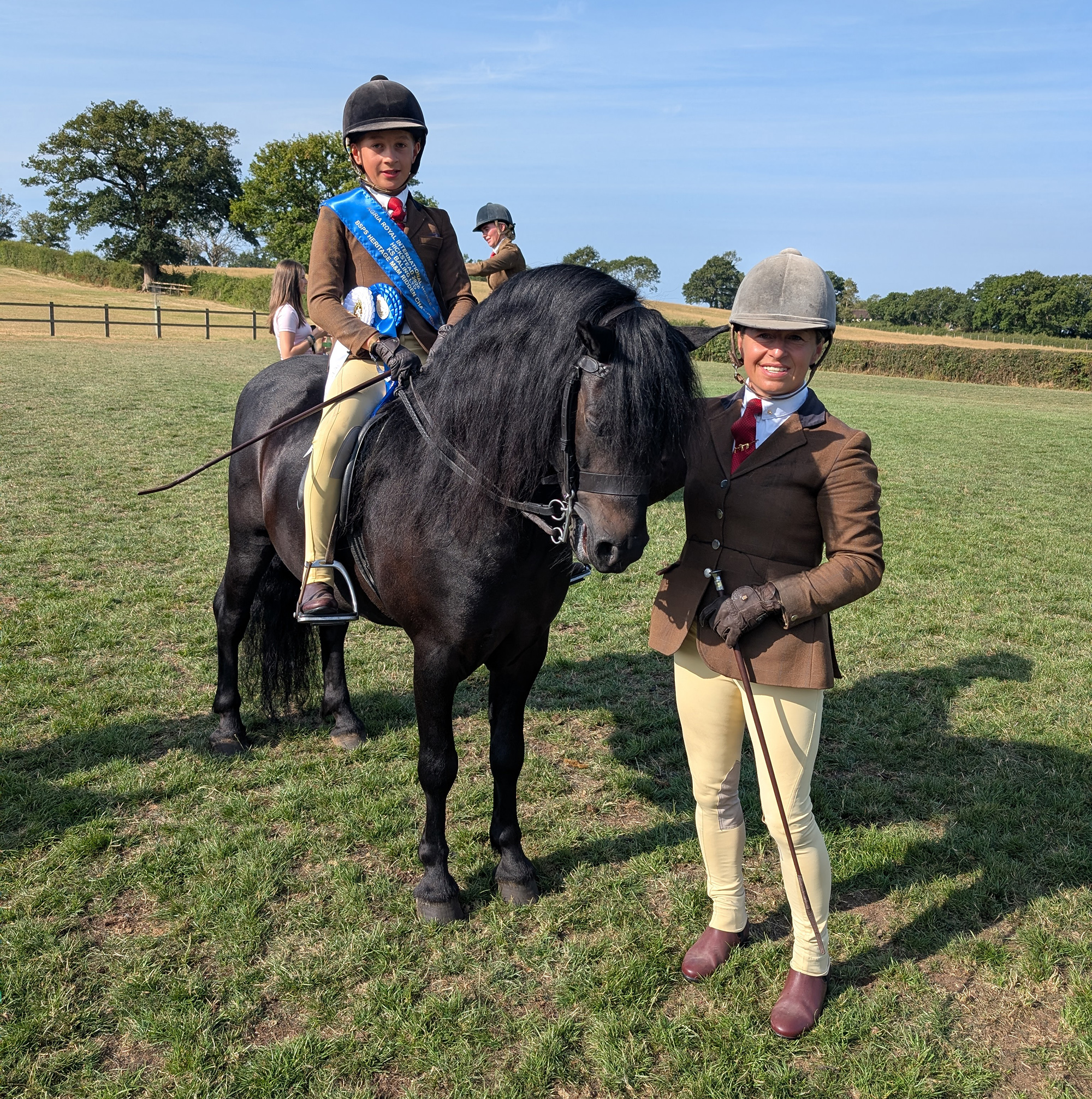 A Dartmoor pony with junior boy rider wearing a blue sash and winners rosettes stood with his mother wearing a tweed showing outfit