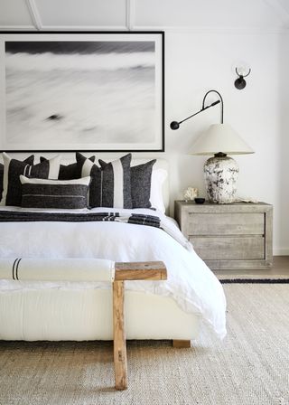 monochrome bedroom with white walls and black charcoal cushions and table lamp with rustic bench at foot of bed