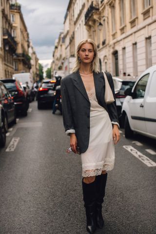 a guest attends paris spring summer menswear fashion week 2025 in a grey blazer, white lace slip dress, black knee high boots, and white shoulder bag