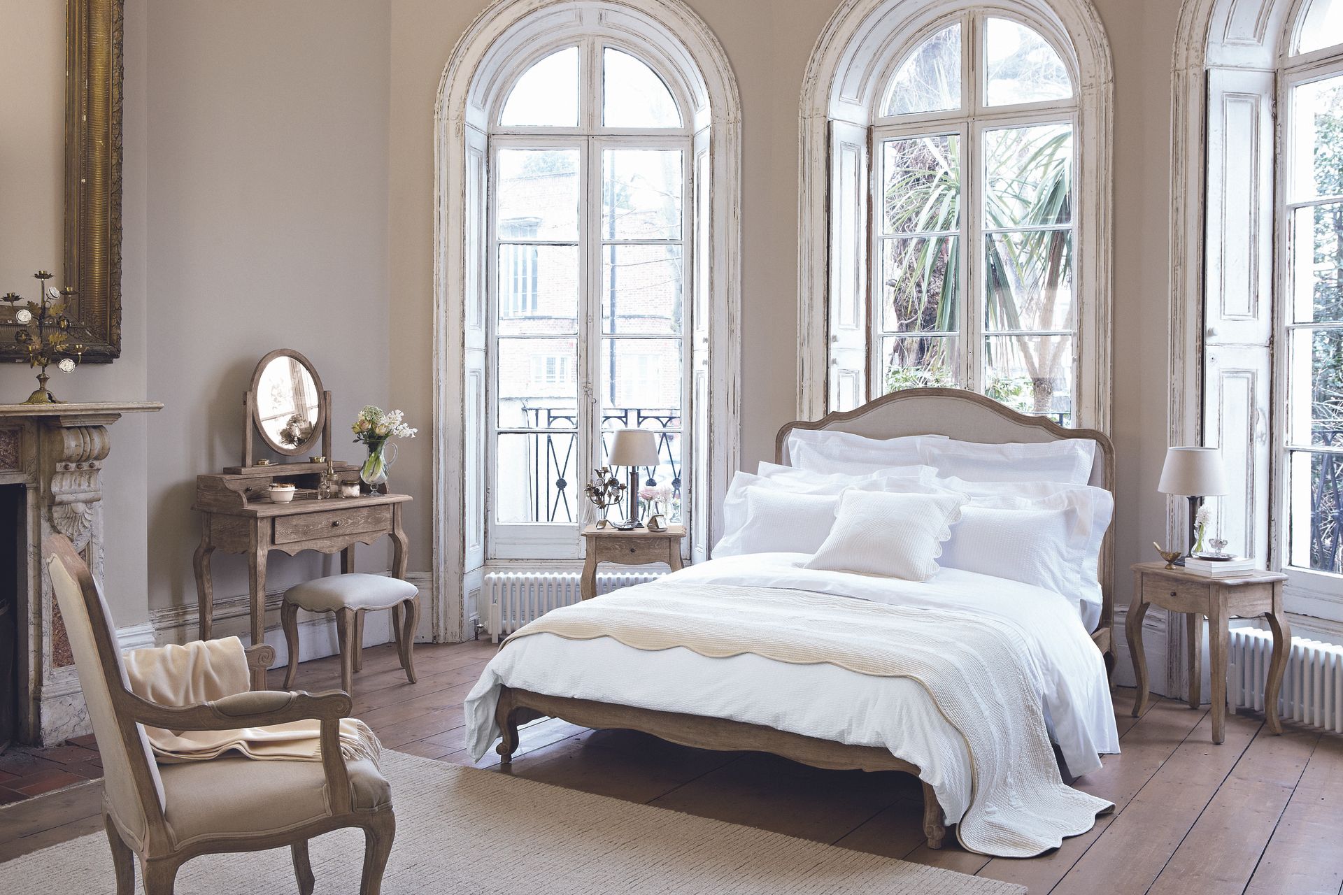 French style bedroom with matching vanity, bed, nightstands and chair. white bedding, vintage gold mirror, bare floorboards, cream rug