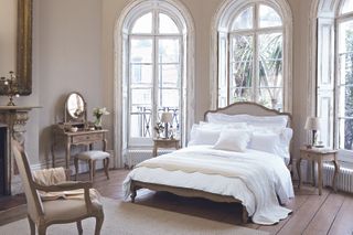 French style bedroom with matching vanity, bed, nightstands and chair. white bedding, vintage gold mirror, bare floorboards, cream rug