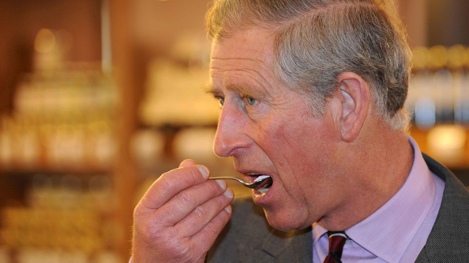 King Charles wears a gray suit and a lilac shirt and eats some food from a spoon