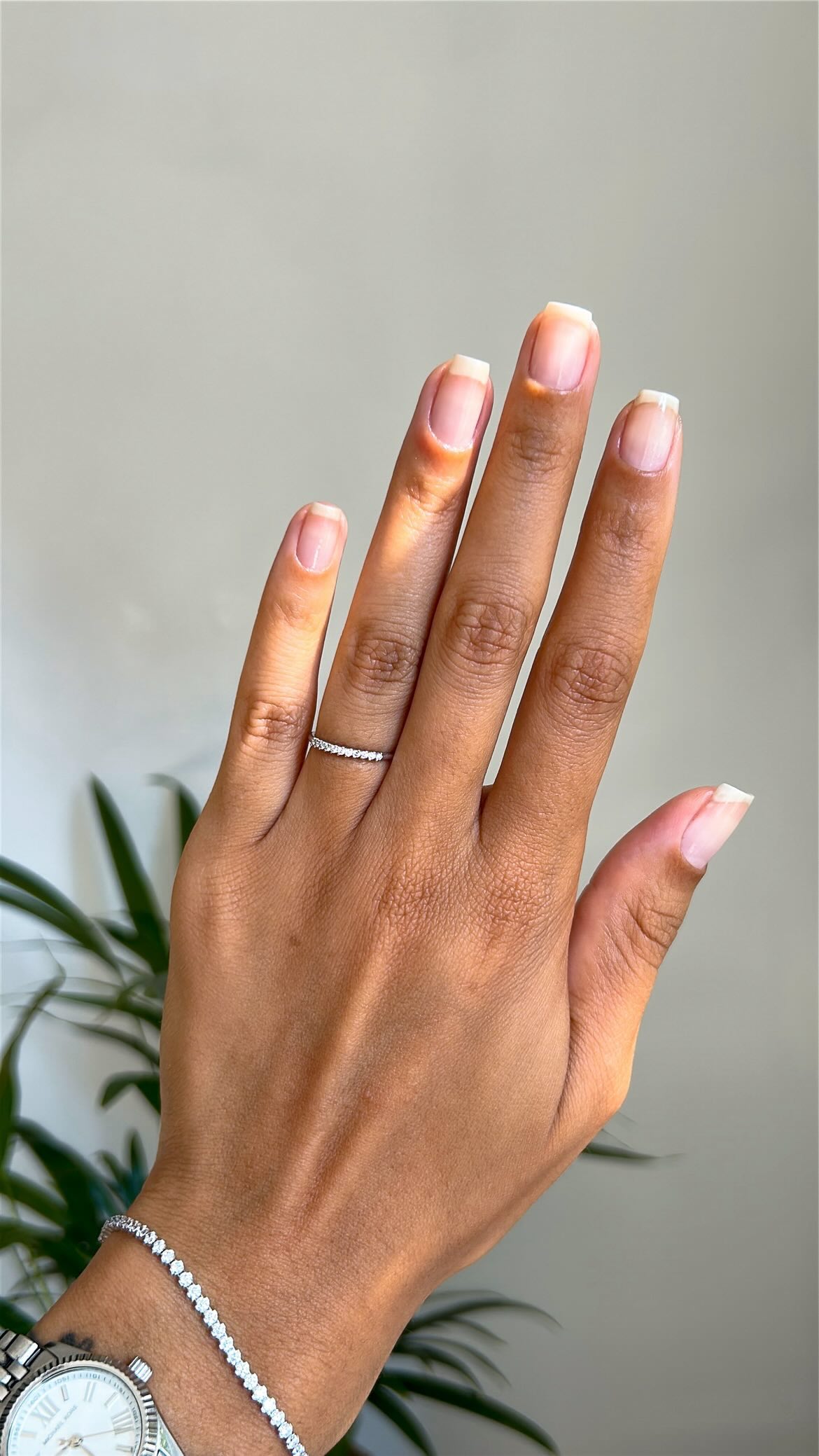 a hand held up in front of a plant with short square, bare nails and a ring on the ring finger as well as a silver tennis bracelet and watch
