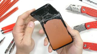 A screenshot from JerryRigEverything's durability test of the OnePlus 10 Pro, showing the phone's damage from a bend test. We see the front of the phone, with the top third bent almost at a right angle and the broken display is lit up in orange below the break line.