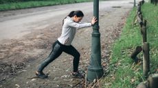woman in leggings and hoodie doing a calf stretch against a lampost in a park setting