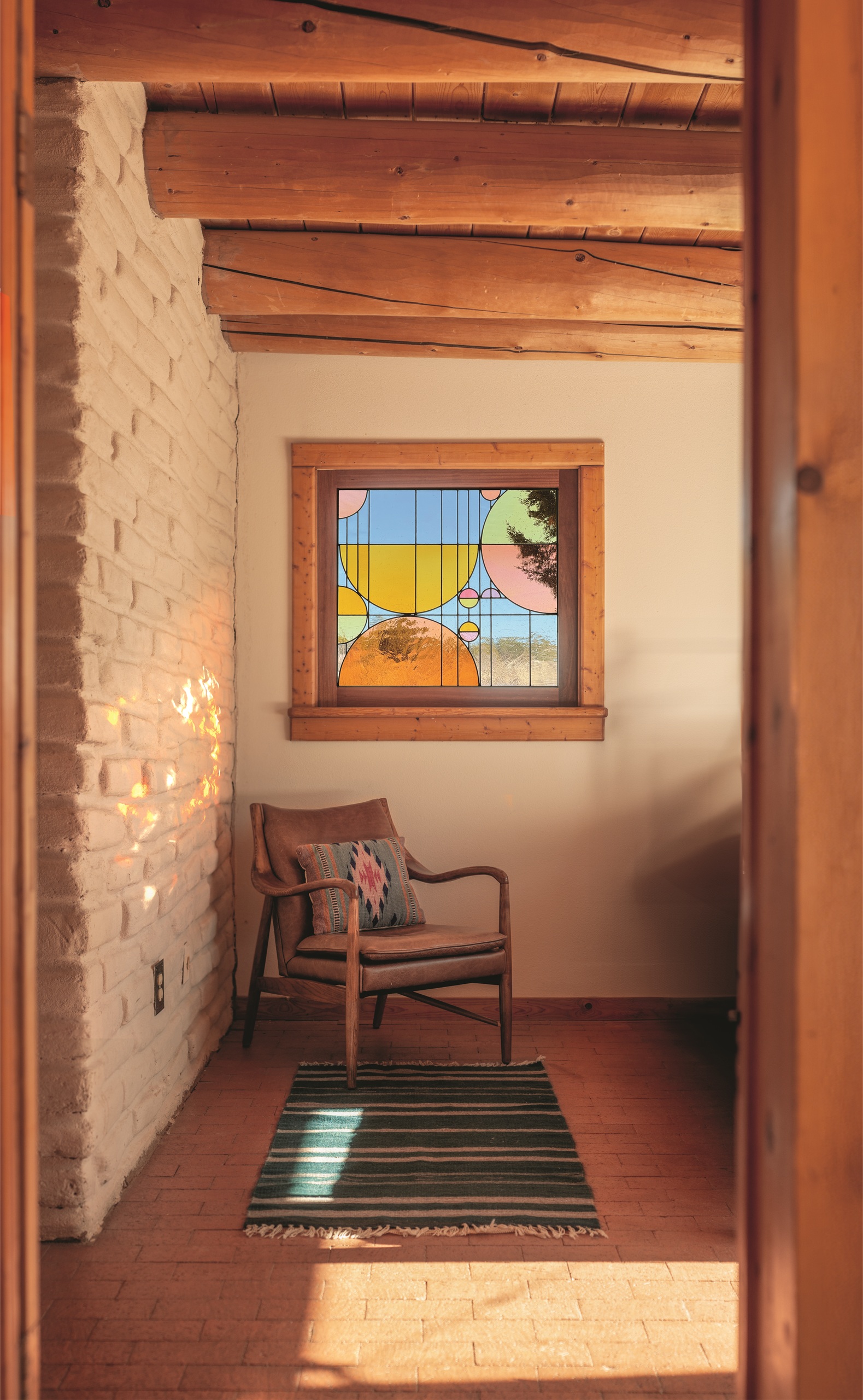room with timber ceiling and exposed beams, brick wall painted white, tiled floor, and armchair and green striped rug under a stained glass window