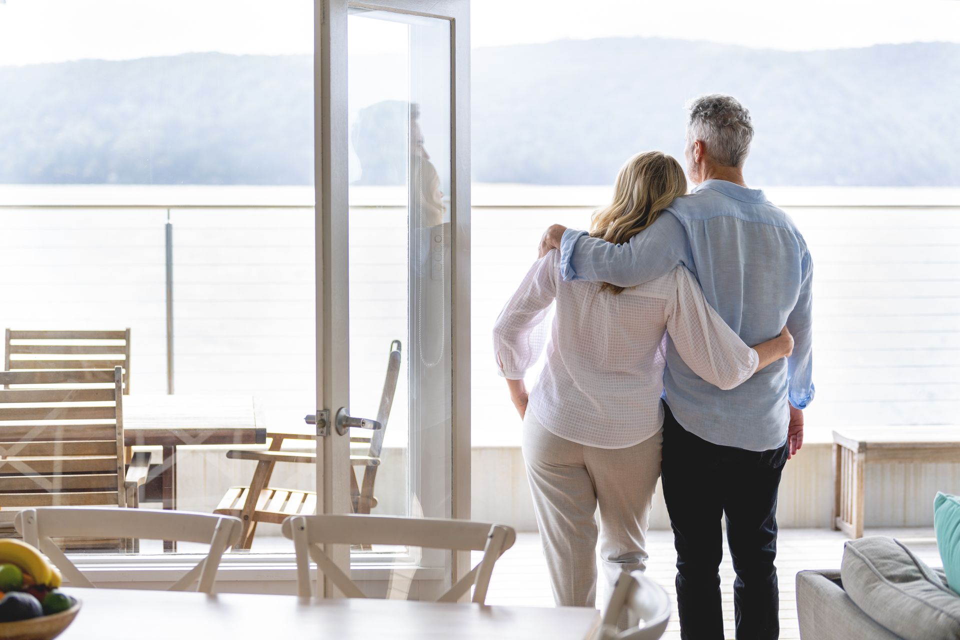 Mature couple looking at the view in their waterfront home. They look happy and contented. They are embracing. The ocean can be seen in the background.