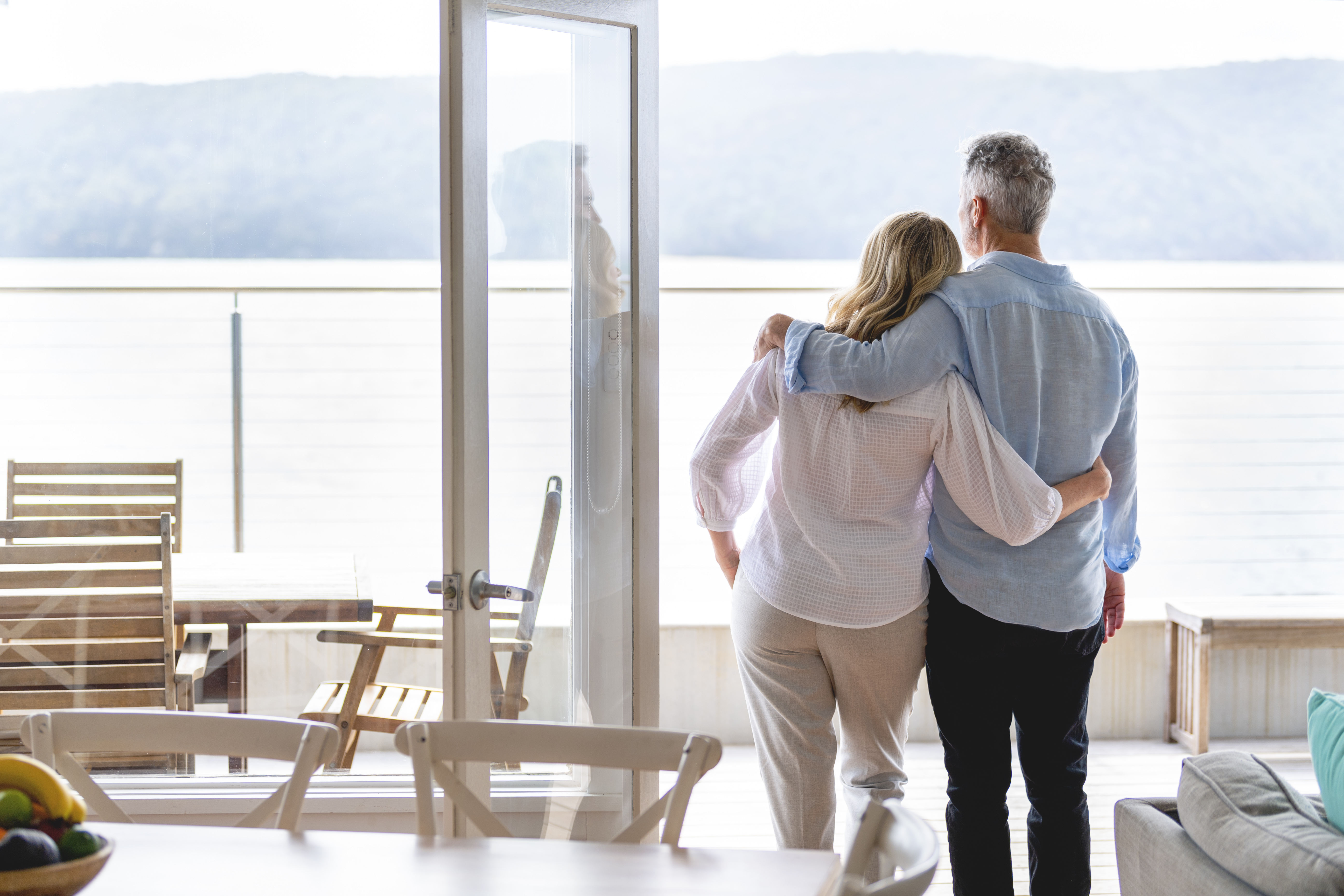 Mature couple looking at the view in their waterfront home. They look happy and contented. They are embracing. The ocean can be seen in the background.