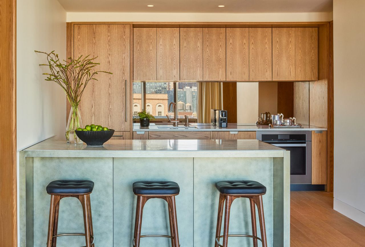 The suite kitchenette of a luxury hotel features desert modernism-inspired furniture in wood and marble, upholstered black leather and wood stools, and plants in a sun-lit environment.