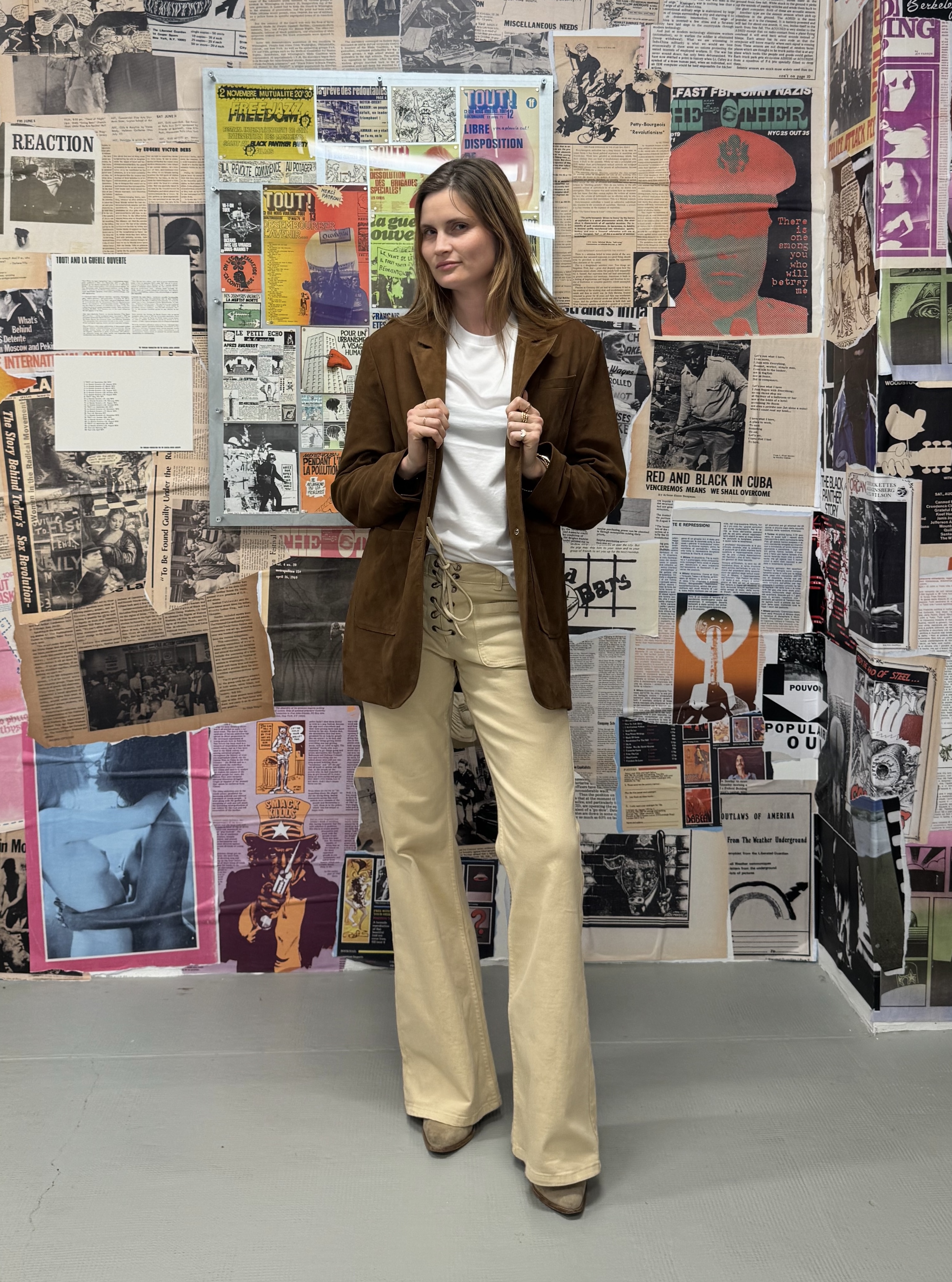 French writer and editor Eugenie Trochu poses in a brown leather blazer, a white T-shirt, lace-up tan flare jeans, and suede boots