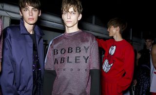 Guys wearing Man S/S 2015 collection. The guy on the left is wearing a navy and purple top with a navy jacket. The guy next to him is wearing a purple top with Bobby Abley London embroidered on it. Next to them is a guy wearing a red tracksuit with a big white smiley face on it.