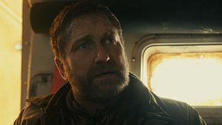 A bearded man (Gerard Butler) looking worried, stood in front a small window.