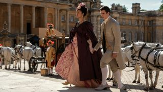 (L to R) India Amarteifio as Young Queen Charlotte waslking up a step while wearing a peach dress and maroon cape with Corey Mylchreest as Young King George.