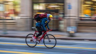 a bicycle courier racing through the streets of a city on a pink singlespeed bike, wearing a yellow beanie style hat