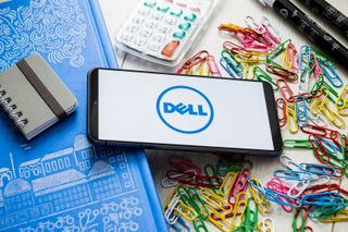 Dell logo on a smartphone, laid down on a desk, one eend slightly leaning on a book which raises the phone. Around it are strewn paperclips, pens and a small calculator