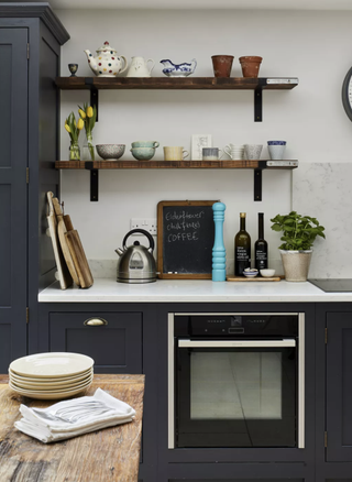 A kitchen with black cabinetry, a countertop with a silver kettle, bottles, and a chalkboard on it, and open shelves with mugs and pots on it