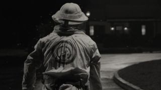 A black and white still of a beekeeper standing in a street wearing a SWORD logo on his back.