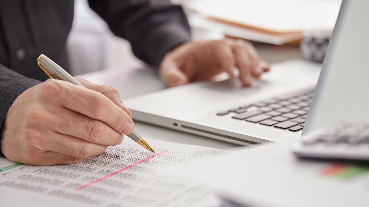 A pair of hands on a desk with one handing resting on the keyboard of a laptop and the other holding a pen above an accounting graphic.