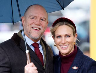 Zara Tindall wearing a blue coat and burgundy turtleneck standing next to Mike Tindall holding an umbrella