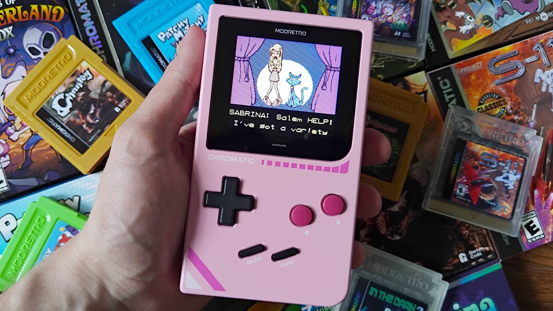 Modretro Chromatic in pink with Game Boy boxes in backdrop and Sabrina Zapped! gameplay on screen.