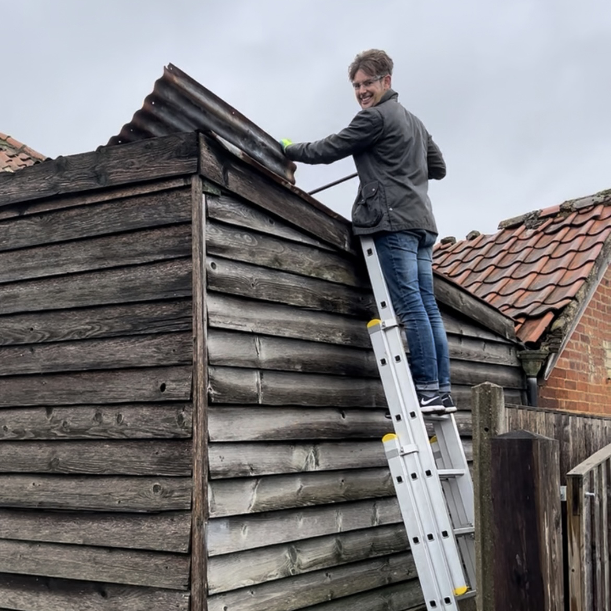 Man stood at the top of a metal extending ladder leant up against a wooden shed with pitched roof