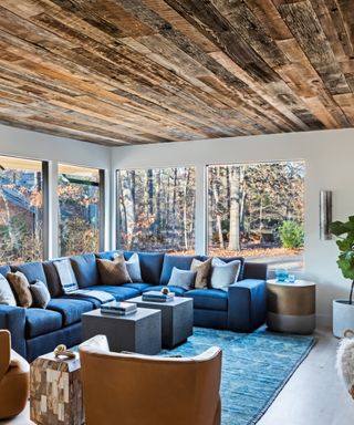 white and blue living room with blue sectional blue rug, tan armchairs and cushions, wooden ceiling, view of outside