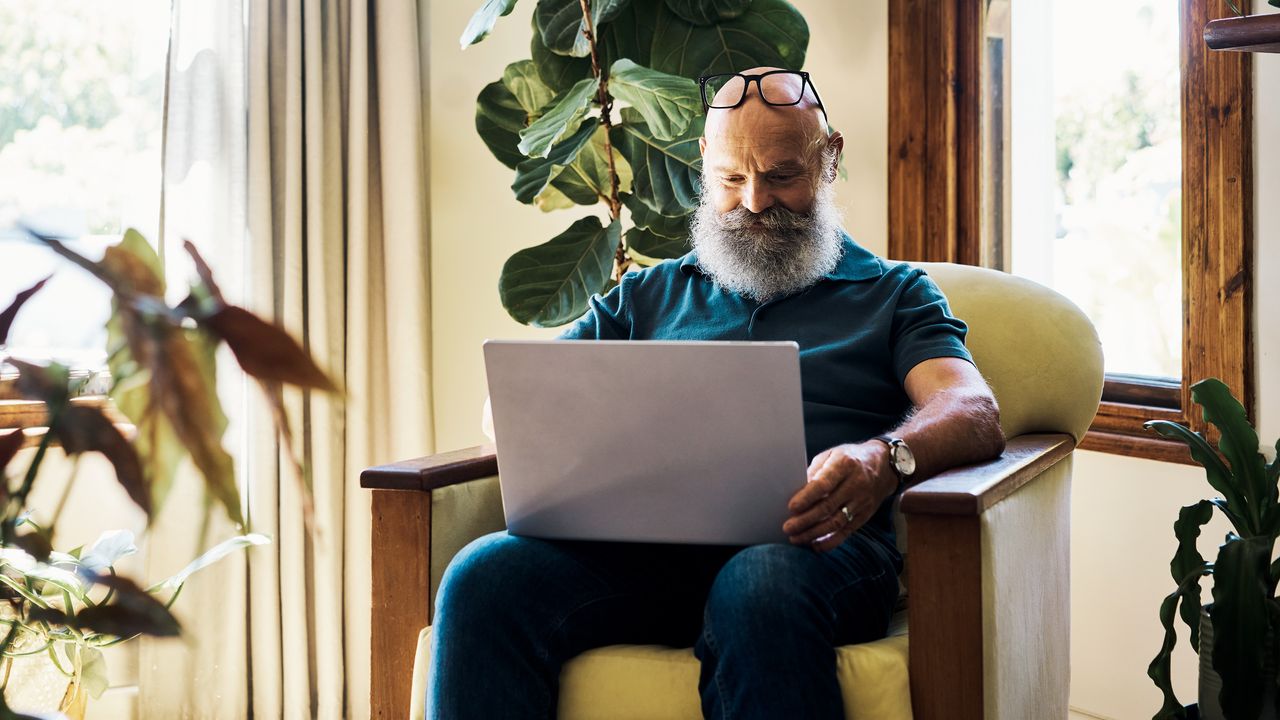 An older man sits in a chair in his living room with an open laptop on his lap.