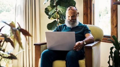 An older man sits in a chair in his living room with an open laptop on his lap.