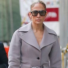 Jennifer Lopez out in New York City wearing a gray belted coat with a brown bag and brown knee-high boots.