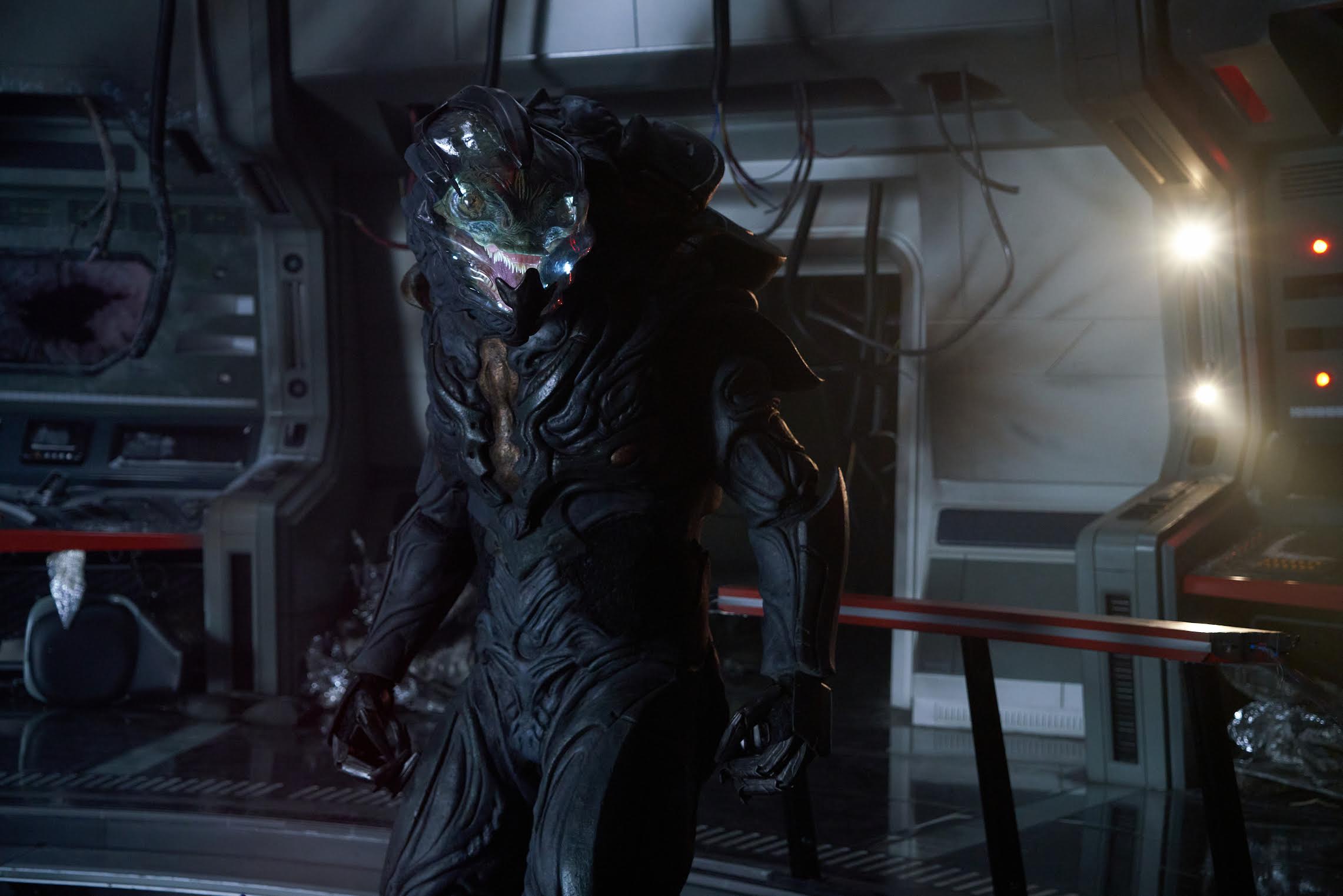 a reptilian alien in a sleek dark spacesuit stands inside a spaceship.
