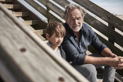 A grandfather and grandson sit on steps near the beach.
