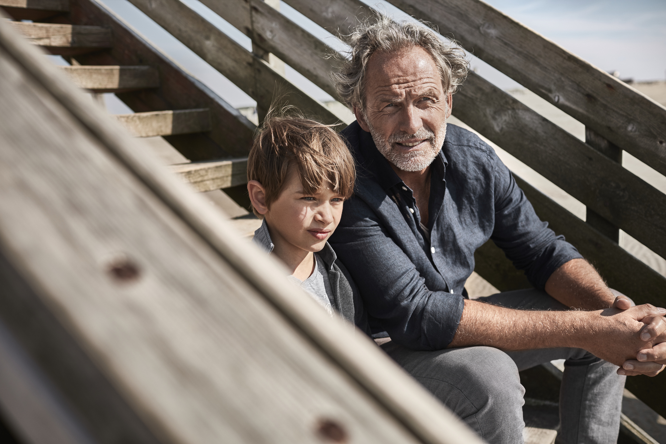 A grandfather and grandson sit on steps near the beach.