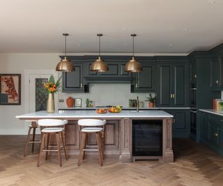 An oak and dark green kitchen with white walls, gold pendant lights, and wood herringbone flooring