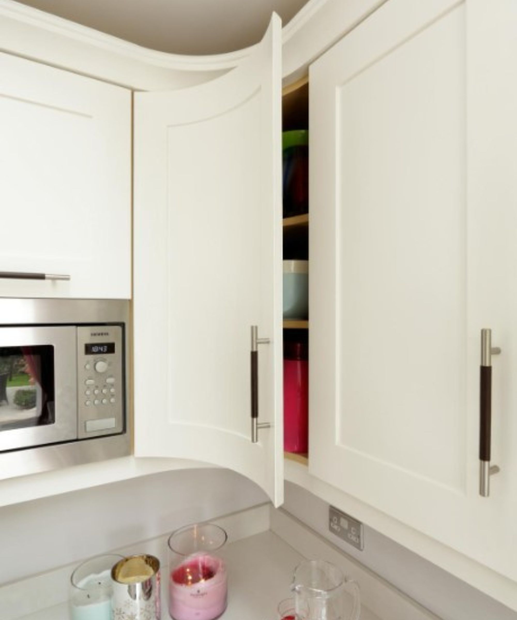 A white curved corner kitchen cabinet that's slightly ajar, with a silver built-in microwave next to it and a white countertop with pink and blue candles underneath it