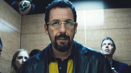 Adam Sandler wearing a black leather jacket, yellow shirt, glasses and earings, in Uncut Gems