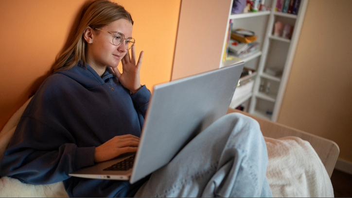 A girl wearing blue hoodie sitting on bed against orange wall studying on laptop