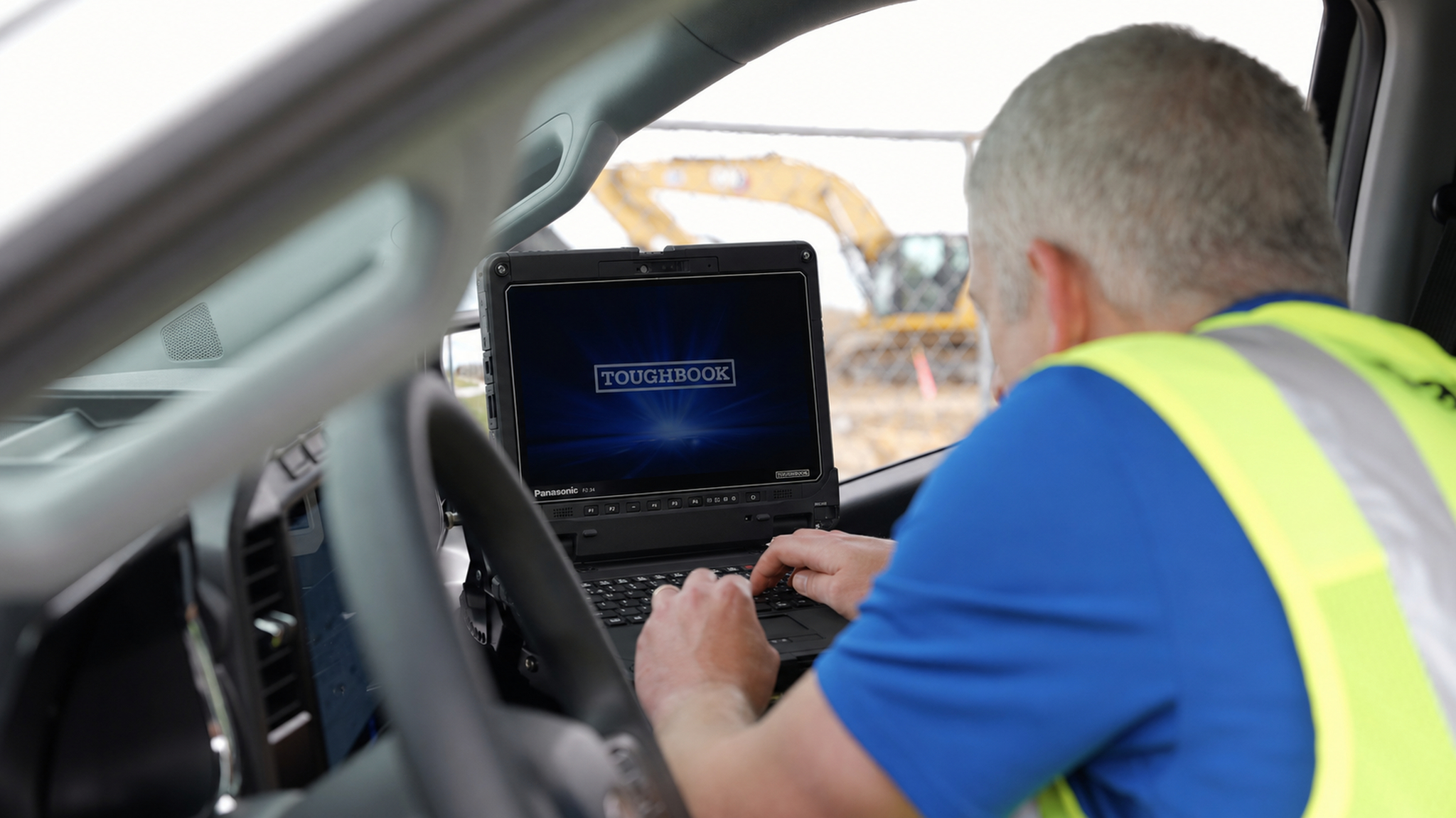 A worker in a high-visibility vest typing on a Panasonic TOUGHBOOK 34 laptop inside a vehicle at a construction site.