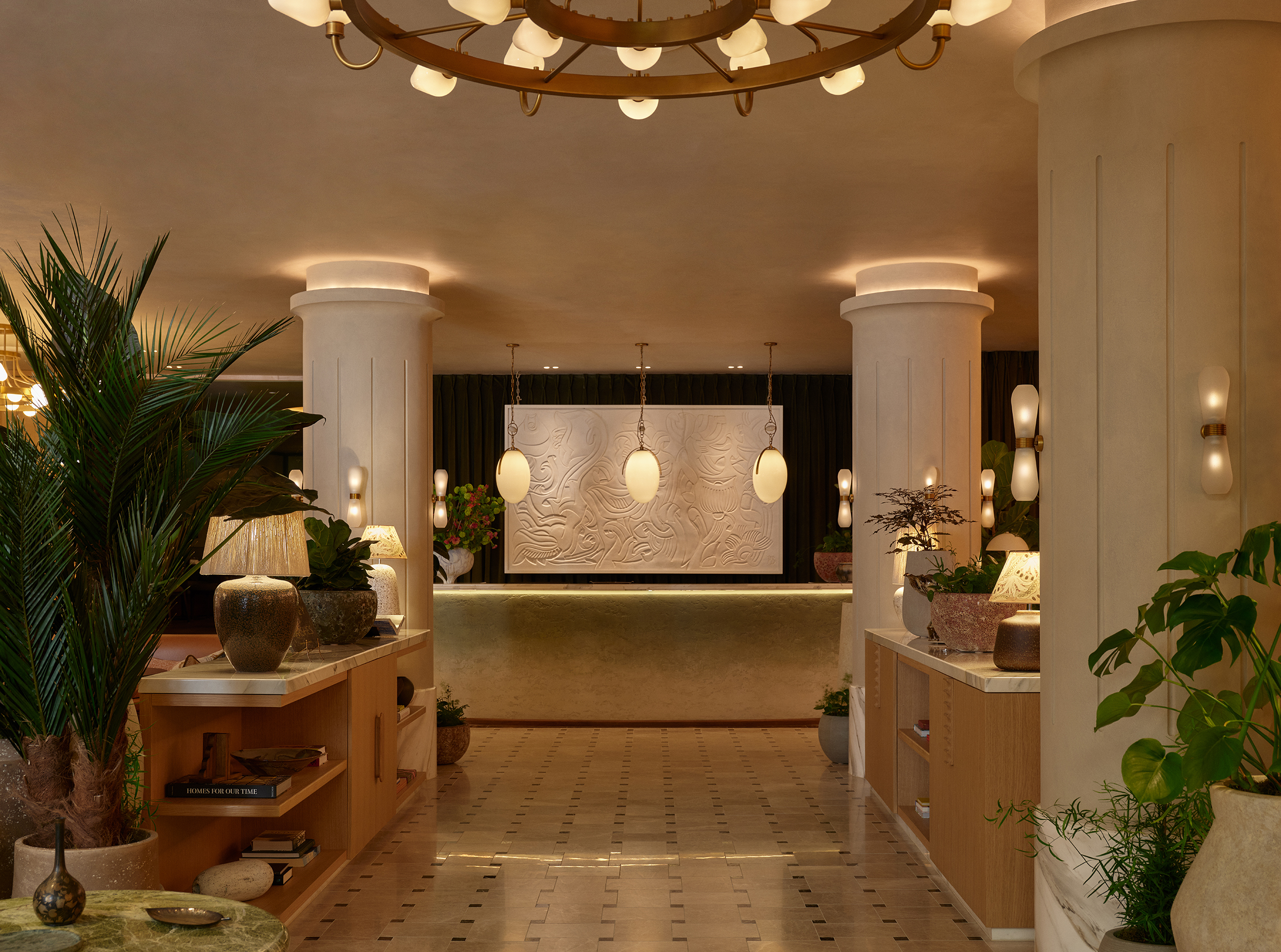 A golden-glowing hotel lobby decorated with smooth wooden cabinetry topped with crafty ceramic lamps and vases, thriving plants, and egg-shaped pendant lights and chandeliers.